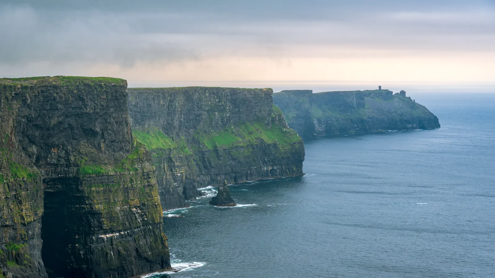 The Cliffs of Moher under a grey sky, the cliff wall running out to O’Brien’s Tower with the sea stack below