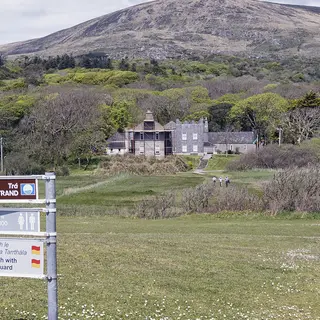 Derrynane House among the trees above the coast, with a walking route signpost in the foreground