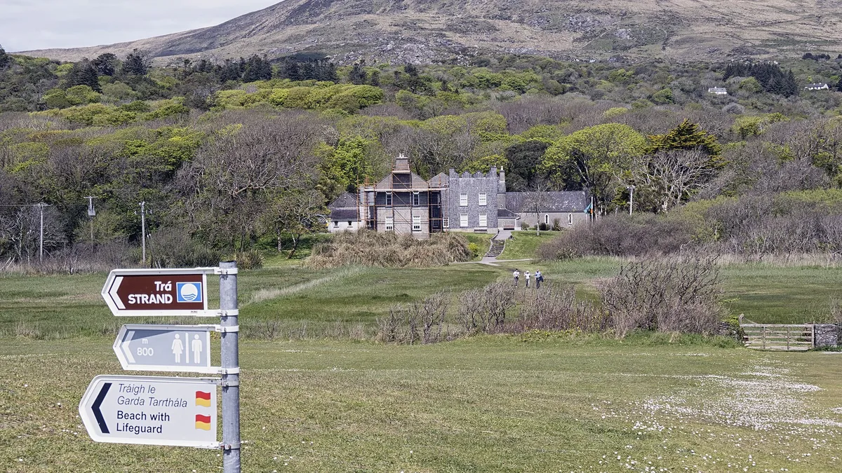 Derrynane House among the trees above the coast, with a walking route signpost in the foreground