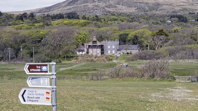 Derrynane House among the trees above the coast, with a walking route signpost in the foreground