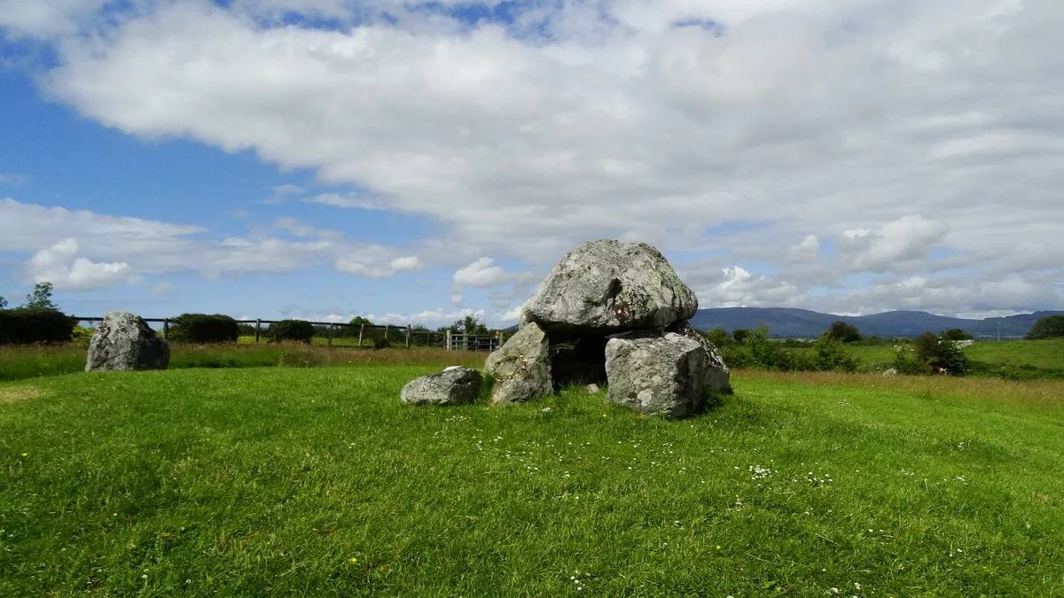 A dolmen of grey boulders in the open field at Carrowmore, with hills beyond